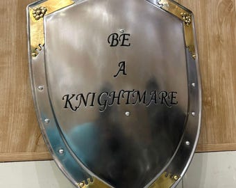 Può includere: Uno scudo color argento con finiture e rivetti color oro. Lo scudo presenta la scritta "BE A KNIGHTMARE" in nero. Lo scudo decorativo è probabilmente per esposizione o costumi, con un tema medievale.