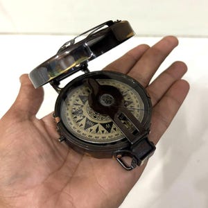 May include: A vintage compass with a hinged lid, held open. The compass has a dark metallic finish with a detailed dial featuring directional markings and degree indicators. The compass is a navigational tool.