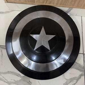 May include: A circular shield in black and silver, featuring a central silver star. The design includes concentric circles of black and silver. This shield is a replica of Captain America's shield.