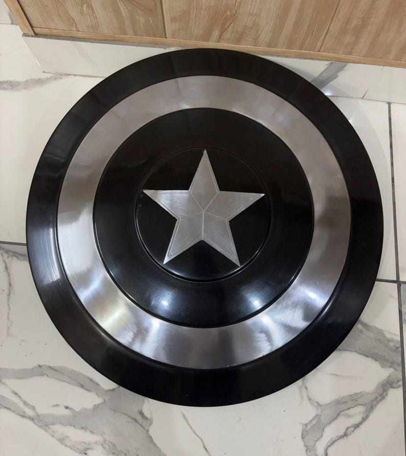May include: A circular shield in black and silver, featuring a central silver star. The design includes concentric circles of black and silver. This shield is a replica of Captain America's shield.
