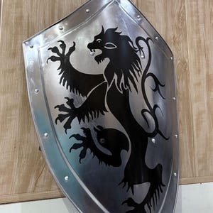 Pu&ograve; includere: Uno scudo color argento con un emblema di leone nero. Lo scudo ha una parte superiore arrotondata ed &egrave; bordato da rivetti. Il leone &egrave; raffigurato in stile araldico, con una zampa alzata e una criniera fluente. Lo scudo &egrave; appoggiato contro una superficie di legno.