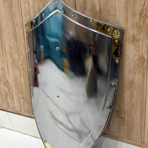 Puede incluir: Un escudo pulido de color plateado, con la parte superior redondeada y la inferior puntiaguda. El escudo tiene un borde dorado y remaches. El escudo est&aacute; apoyado contra una pared de madera. Es un objeto decorativo.
