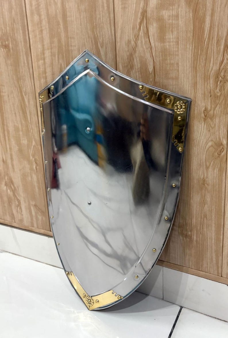 Puede incluir: Un escudo pulido de color plateado, con la parte superior redondeada y la inferior puntiaguda. El escudo tiene un borde dorado y remaches. El escudo est&aacute; apoyado contra una pared de madera. Es un objeto decorativo.