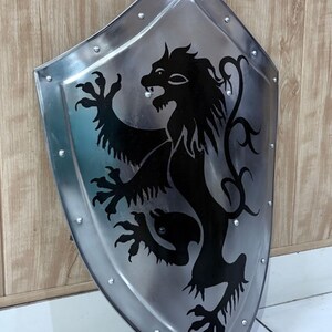 Pu&ograve; includere: Scudo color argento con un emblema di leone nero. Lo scudo &egrave; rivettato sui bordi e ha una base appuntita. Il leone &egrave; raffigurato in stile araldico, con un'espressione feroce e le zampe sollevate. Lo scudo &egrave; probabilmente in metallo.