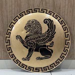 May include: A circular gold-toned shield featuring a prominent black graphic of a winged lion in the center. The outer edge of the shield is decorated with a continuous black Greek key pattern, creating a bold contrast against the metallic surface.