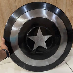 May include: A black and silver shield with a central silver star design. The shield has a circular shape with concentric rings of black and silver. The shield is held by a person's hand.