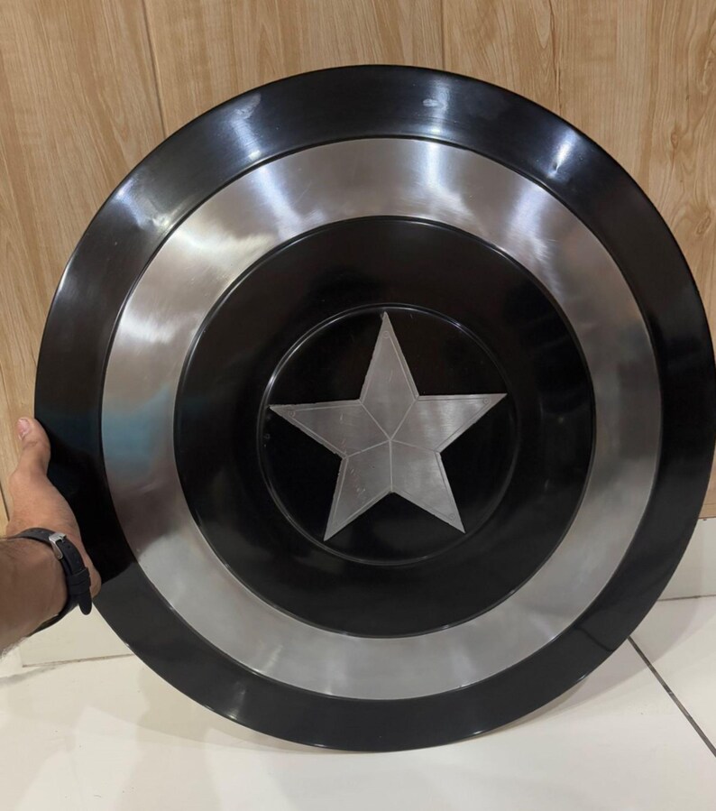 May include: A black and silver shield with a central silver star design. The shield has a circular shape with concentric rings of black and silver. The shield is held by a person's hand.