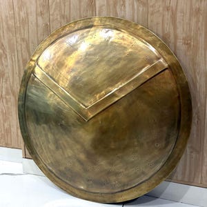 Op de afbeelding: Een groot, rond, convex schild gemaakt van gepolijst messing met een goudbruine metallic afwerking. Het oppervlak is voorzien van een opvallende V-vormige verhoogde rand in het midden en talloze kleine, gelijkmatig verdeelde cirkelvormige inkepingen.