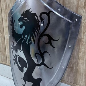 Pu&ograve; includere: Uno scudo color argento con un emblema di leone nero. Lo scudo ha una parte superiore arrotondata e una inferiore a punta, con rivetti sul bordo. Il leone &egrave; raffigurato in stile araldico.