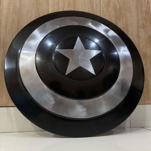 May include: A black and silver shield featuring a central silver star. The shield has a circular design with concentric rings of black and silver. A five-pointed star is in the center. The shield is a prop or collectible.