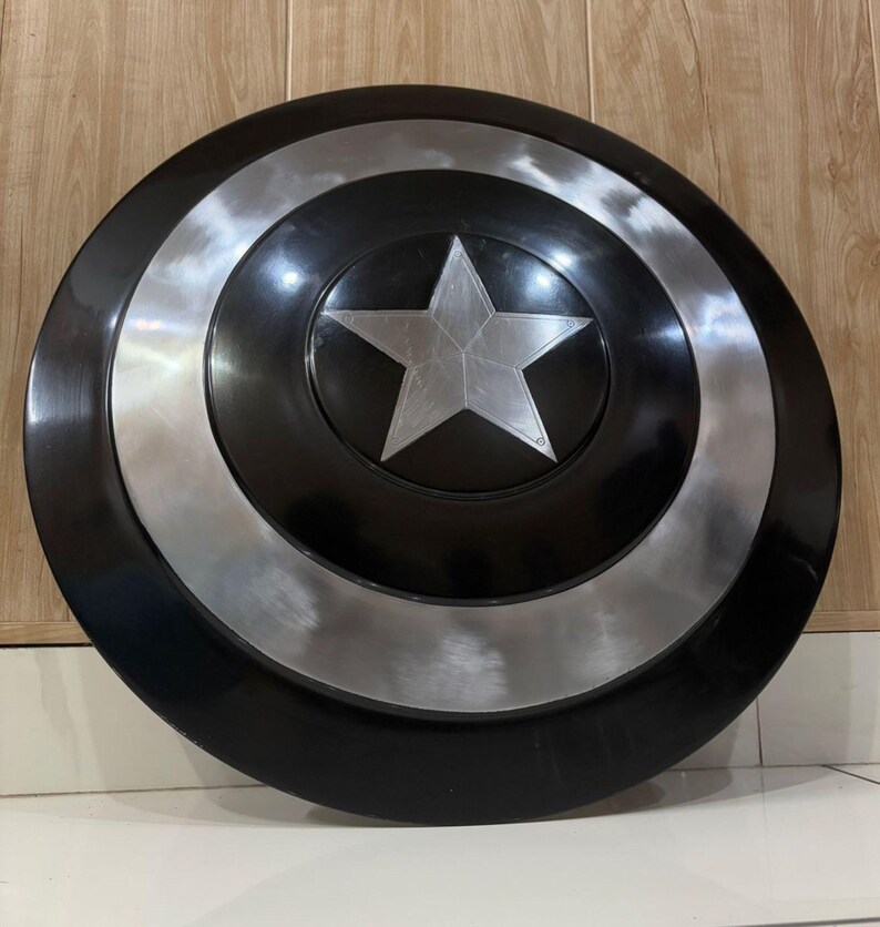 May include: A black and silver shield featuring a central silver star. The shield has a circular design with concentric rings of black and silver. A five-pointed star is in the center. The shield is a prop or collectible.