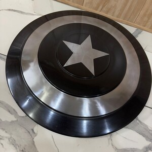 May include: A black and silver Captain America shield. The shield features a central silver star on a black base, encircled by silver and black rings. The round shield is metallic in appearance.