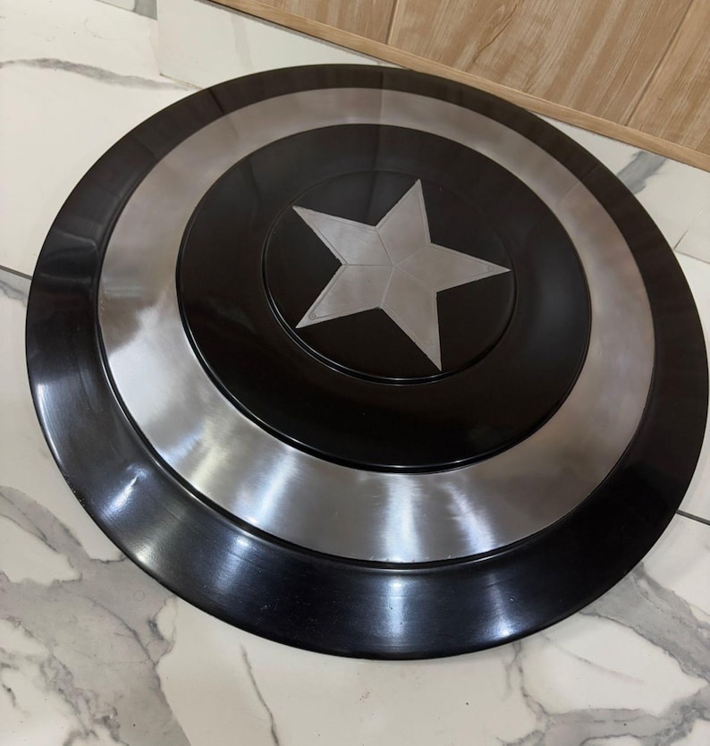 May include: A black and silver Captain America shield. The shield features a central silver star on a black base, encircled by silver and black rings. The round shield is metallic in appearance.