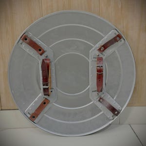 May include: A silver-coloured, circular shield with brown leather straps and buckles on the back. The shield has concentric circular ridges and riveted metal supports for the straps. The leather straps are secured with metal rivets.