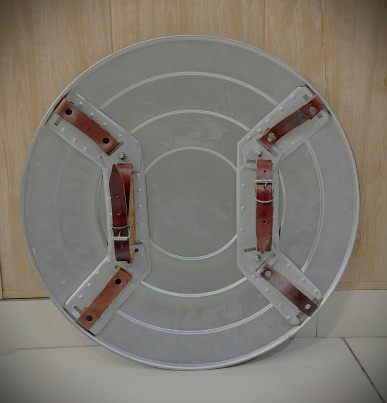 May include: A silver-coloured, circular shield with brown leather straps and buckles on the back. The shield has concentric circular ridges and riveted metal supports for the straps. The leather straps are secured with metal rivets.