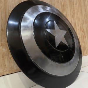 May include: A black and silver shield featuring a large silver star at its centre. The shield has concentric black and silver circles. This shield is a replica of Captain America's shield.