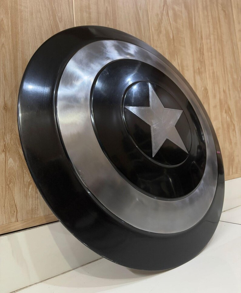 May include: A black and silver shield featuring a large silver star at its centre. The shield has concentric black and silver circles. This shield is a replica of Captain America's shield.