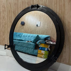 Może przedstawiać: Okrągłe, czarne, metalowe lustro z motywem iluminatora. Lustro odbija turkusową sofę i drewniane panele. Rama posiada nity i uchwyt, co dodaje mu morskiego charakteru. To dekoracja ścienna.