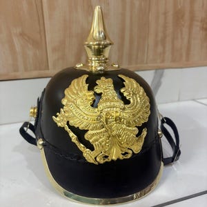 May include: A black helmet with a gold-colored eagle emblem and a gold spike on top. The helmet has a gold trim around the base and a chin strap. The eagle emblem is detailed with outstretched wings.