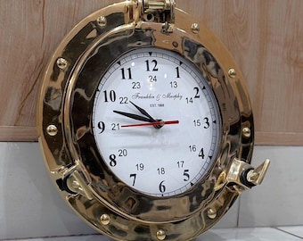 May include: A circular wall clock with a polished brass porthole frame featuring a white face, black hour and minute hands, and a red second hand. The dial displays numbers 1 through 12 and 13 through 24, with the text Franklin & Murphy EST. 1868.