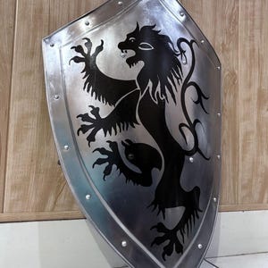 Pu&ograve; includere: Uno scudo color argento con un emblema di leone nero. Lo scudo ha una parte superiore arrotondata e una inferiore a punta, con rivetti lungo i bordi. Il leone &egrave; raffigurato in stile araldico.