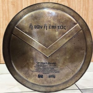 Op de afbeelding: Een cirkelvormig, bronskleurig schild met een verhoogd V-vormig embleem in het midden. De Griekse tekst "ἢ τὰν ἢ ἐπὶ τᾶς" is boven het embleem gegraveerd, terwijl de naam "CPT Paul J. Murphy" en een herdenkingstekst eronder staan.