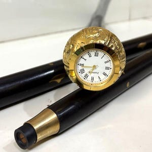 May include: A black wooden walking cane with gold accents and a decorative gold watch. The watch has a white face with black Roman numerals and the word "QUARTZ" at the bottom. The cane is a vintage style.