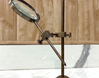 May include: A vintage-style brass magnifying glass on a stand. The magnifying glass is round with a clear lens and a brass frame. The stand has a round base, a vertical pole, and an adjustable arm to hold the magnifying glass.