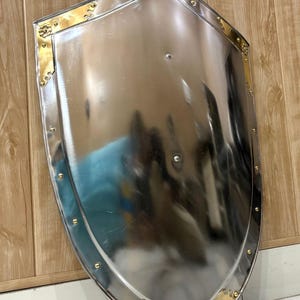 Puede incluir: Un escudo pulido de color plateado, con la parte superior redondeada y la inferior puntiaguda. El escudo tiene detalles decorativos dorados en las esquinas y a lo largo de los bordes, con peque&ntilde;os remaches. El escudo se muestra sobre un fondo de madera.