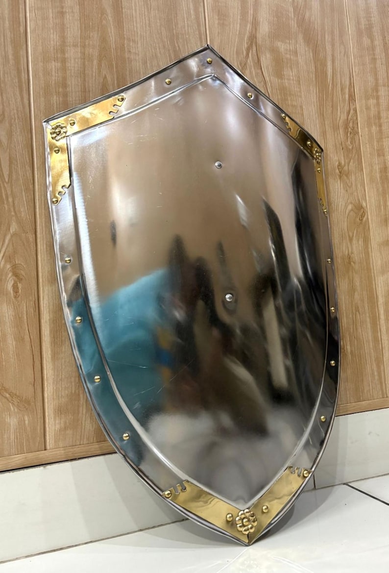 Puede incluir: Un escudo pulido de color plateado, con la parte superior redondeada y la inferior puntiaguda. El escudo tiene detalles decorativos dorados en las esquinas y a lo largo de los bordes, con peque&ntilde;os remaches. El escudo se muestra sobre un fondo de madera.