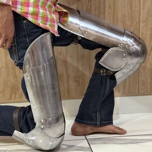 May include: A pair of polished steel leg armor pieces consisting of articulated greaves and poleyns. The metal armor features visible rivets and brown leather straps with metal buckles, worn over dark blue denim jeans and a plaid shirt.