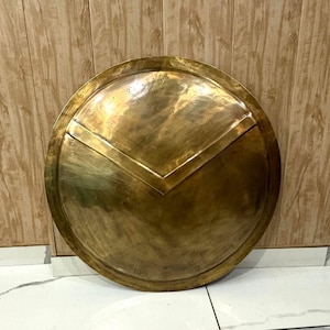 Op de afbeelding: Een grote, ronde schild met een messing afwerking, in antieke stijl. Het heeft een opvallend V-vormig ontwerp in het midden en kleine, decoratieve klinknagels. Het uiterlijk suggereert een historisch of decoratief doel, met een diameter van ongeveer 76 cm.