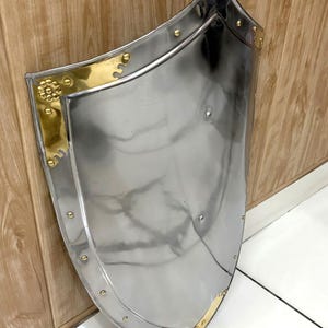 Puede incluir: Un escudo pulido de color plateado con detalles en lat&oacute;n. El escudo tiene una forma curva, similar a una cometa, con esquinas y remaches de lat&oacute;n decorativos. Los bordes est&aacute;n adornados con lat&oacute;n. El escudo est&aacute; apoyado contra una superficie de madera.