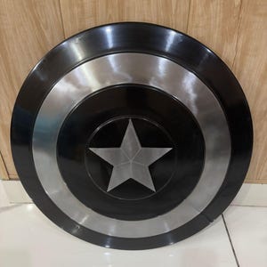 May include: A circular shield featuring a black and silver design. The shield has a central silver star and concentric circles in black and silver. This shield is a prop or collectible item.