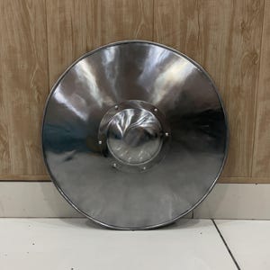 May include: A circular metal shield with a polished silver finish. The shield features a prominent central dome, known as a boss, which is securely attached to the main body with six visible rivets arranged in a symmetrical pattern around the center.