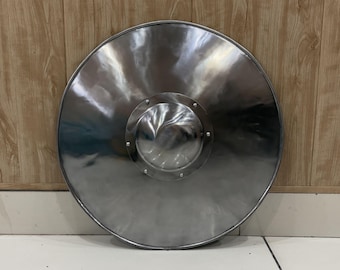 Può includere: Uno scudo metallico circolare con una finitura argento lucido. Lo scudo presenta una cupola centrale prominente, nota come umbone, che è fissata saldamente al corpo principale con sei rivetti visibili disposti in uno schema simmetrico.