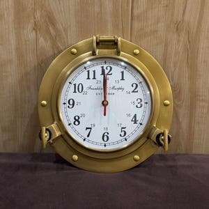 Può includere: Un orologio da parete color ottone con quadrante bianco e numeri neri. L'orologio ha un design a oblò nautico con una parte superiore incernierata e chiusure laterali. Il quadrante dell'orologio recita "Franklin Murphy EST 1868".