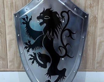 Puede incluir: Un escudo plateado con un emblema de león negro. El escudo tiene una base puntiaguda y está bordeado con remaches. El león está en una pose heráldica, con sus patas delanteras levantadas. El escudo es probablemente de metal.