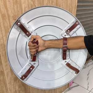 May include: A silver-coloured, round shield with a dark brown leather handle and arm straps. The shield has a star-shaped design with four brown leather straps attached to the back for grip and arm support. The shield is made of metal.