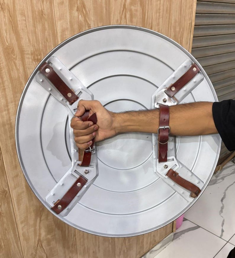 May include: A silver-coloured, round shield with a dark brown leather handle and arm straps. The shield has a star-shaped design with four brown leather straps attached to the back for grip and arm support. The shield is made of metal.