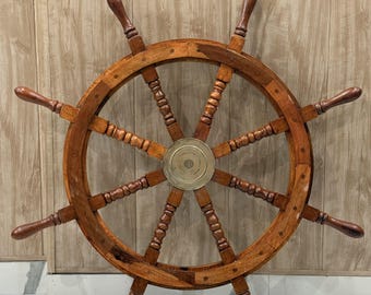 May include: A large wooden ship's wheel featuring eight spokes with rounded handles and a central circular brass hub. The wheel is constructed from polished brown wood with visible grain patterns, showcasing a classic nautical design element.