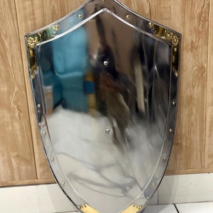 Puede incluir: Un escudo plateado pulido con un borde dorado y remaches decorativos. El escudo tiene un dise&ntilde;o medieval cl&aacute;sico, con una superficie ligeramente curvada y una parte inferior puntiaguda. El borde dorado presenta detalles ornamentados.