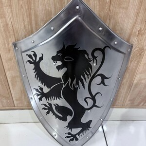 Pu&ograve; includere: Uno scudo color argento con un emblema di leone nero. Lo scudo ha una parte superiore arrotondata e una inferiore a punta, con rivetti lungo i bordi. Il leone &egrave; raffigurato in stile araldico.