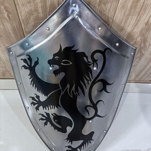 Pu&ograve; includere: Uno scudo in metallo color argento con un emblema di leone nero. Lo scudo ha una parte superiore a punta e una base arrotondata, con rivetti lungo i bordi. Il leone &egrave; raffigurato in stile araldico, con una zampa alzata e una criniera fluente.