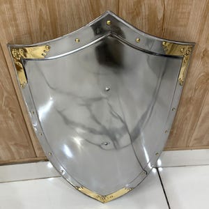 Puede incluir: Un escudo plateado pulido con una parte superior curvada y una base puntiaguda. El escudo presenta detalles dorados a lo largo de los bordes y est&aacute; remachado con peque&ntilde;os pernos redondos. El escudo es probablemente de metal y est&aacute; dise&ntilde;ado para la protecci&oacute;n.