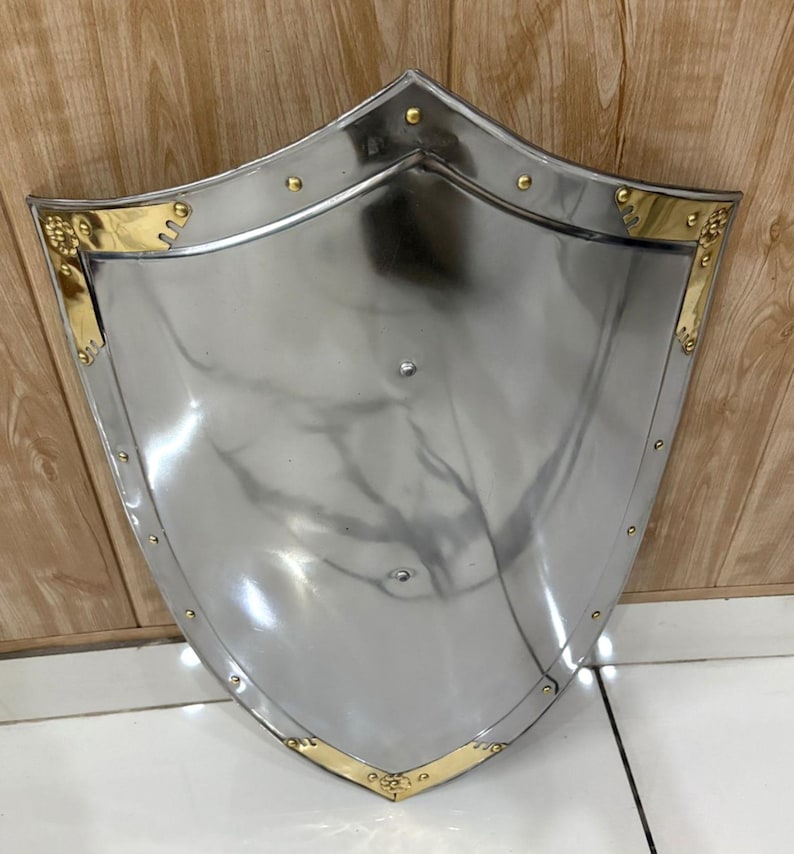 Puede incluir: Un escudo plateado pulido con una parte superior curvada y una base puntiaguda. El escudo presenta detalles dorados a lo largo de los bordes y est&aacute; remachado con peque&ntilde;os pernos redondos. El escudo es probablemente de metal y est&aacute; dise&ntilde;ado para la protecci&oacute;n.