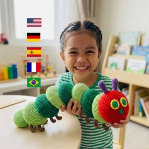 May include: A handmade, crocheted caterpillar toy in shades of green and red. The caterpillar has a smiling face with yellow and green accents. The toy is held by a smiling child. The caterpillar is approximately 12 inches long.