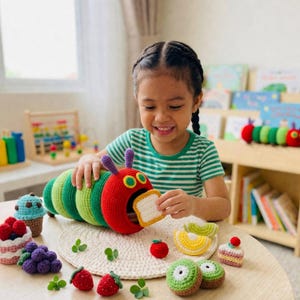 May include: A crocheted caterpillar toy with a red head and green and yellow body, open mouth holding a piece of bread. Surrounding the caterpillar are crocheted fruits and desserts, including strawberries, kiwi, and cupcakes. The toys are on a round table.