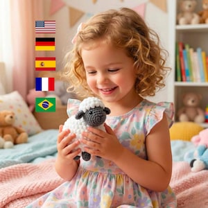 Può includere: Una bambina sorride mentre tiene in mano un piccolo giocattolo a forma di pecora fatto all'uncinetto. La pecora ha un corpo bianco, testa e orecchie grigie e occhi neri. La bambina indossa un vestito floreale.