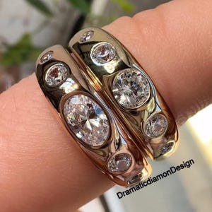 Puede incluir: Dos anillos de oro con múltiples piedras preciosas claras y brillantes. Un anillo presenta una piedra central de forma ovalada, mientras que el otro tiene una piedra central redonda. Los anillos están engastados con piedras más pequeñas. Los anillos están en un dedo.